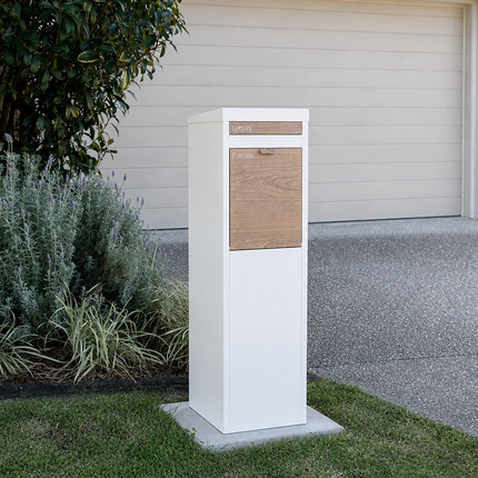 A modern white outdoor mailbox with a natural wood front panel and a small mail slot, mounted on a concrete base in a grassy lawn next to a house driveway.