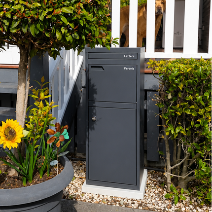 A dark gray outdoor mailbox positioned next to a large gray planter holding a trimmed topiary tree and bright yellow sunflowers, set against a white-fronted house with steps.
