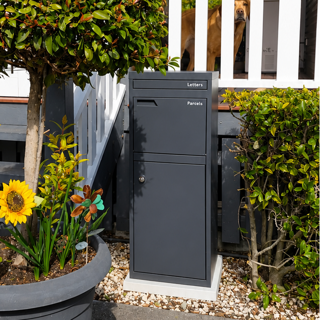 A dark gray outdoor mailbox positioned next to a large gray planter holding a trimmed topiary tree and bright yellow sunflowers, set against a white-fronted house with steps.