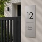 A modern rectangular brushed-metal house number sign reading '12' with 'Collins Street' below, mounted on a light stucco wall beside a black gate and greenery.